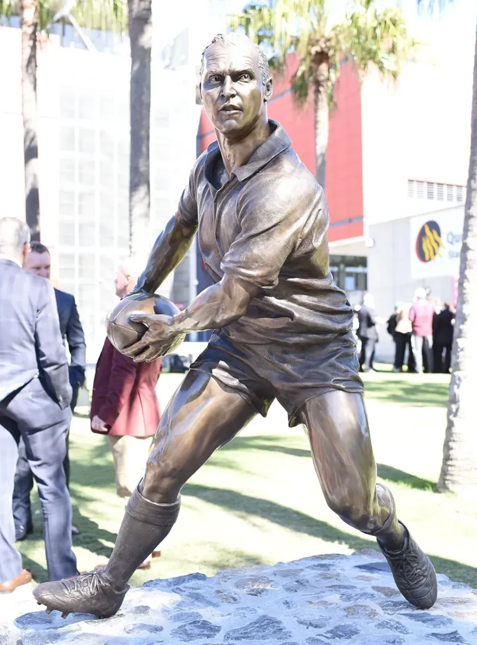 Bespoke Bronze Memorial of a rugby player passing a ball, displayed outdoors on a stone pedestal with crowd and modern buildings in the background.