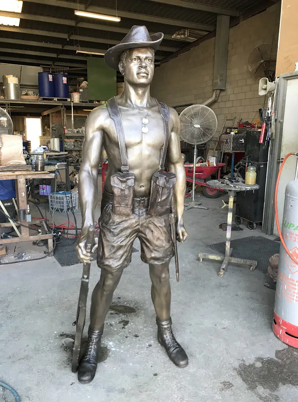 Bespoke Bronze Memorial statue of a smiling, muscular man with hat, dog tags, and rifle, handcrafted in a workshop; ideal custom memorial sculpture.