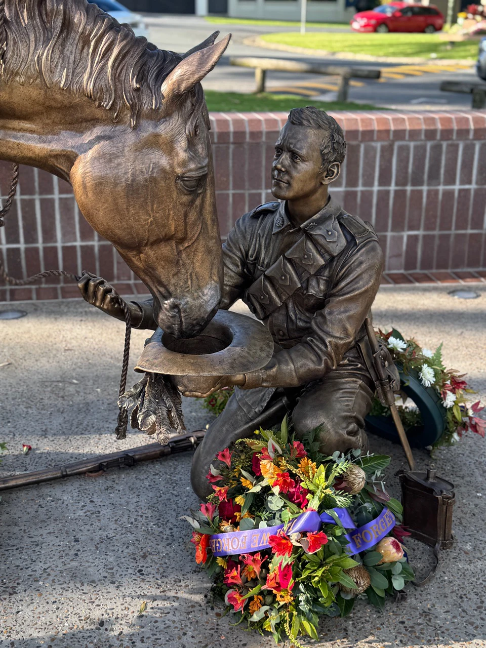 Bespoke Bronze Memorial: Bronze statue of a soldier kneeling, offering water to a horse with his hat, surrounded by floral wreaths and a brick wall backdrop.