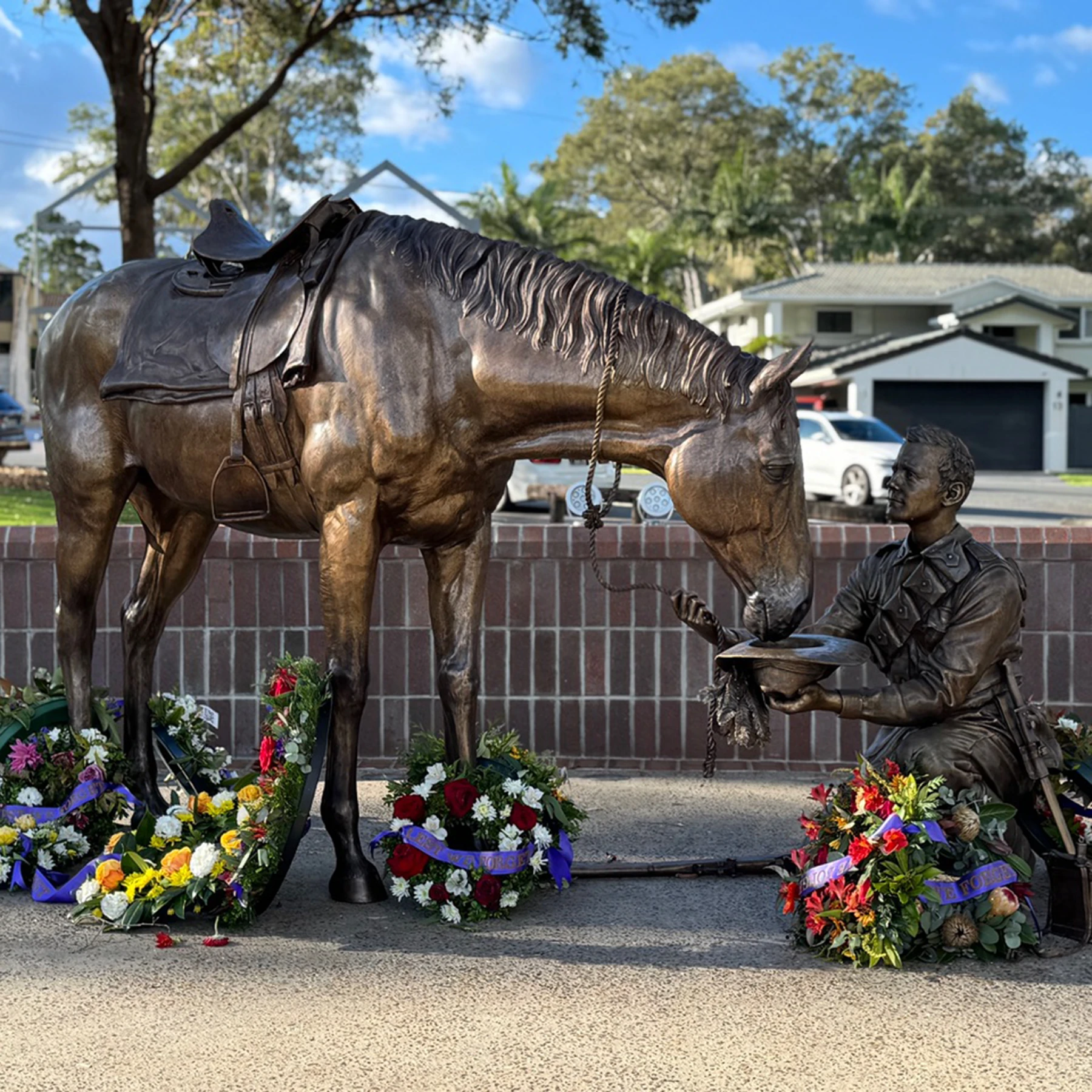 Bespoke Bronze Memorial statue of a kneeling soldier giving water to a horse, surrounded by wreaths, outdoors near brick wall and suburban homes.