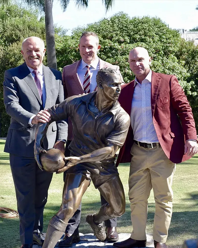 Three men in suits smile by a Bespoke Bronze Memorial rugby player statue outdoors, with trees and a building in the background.